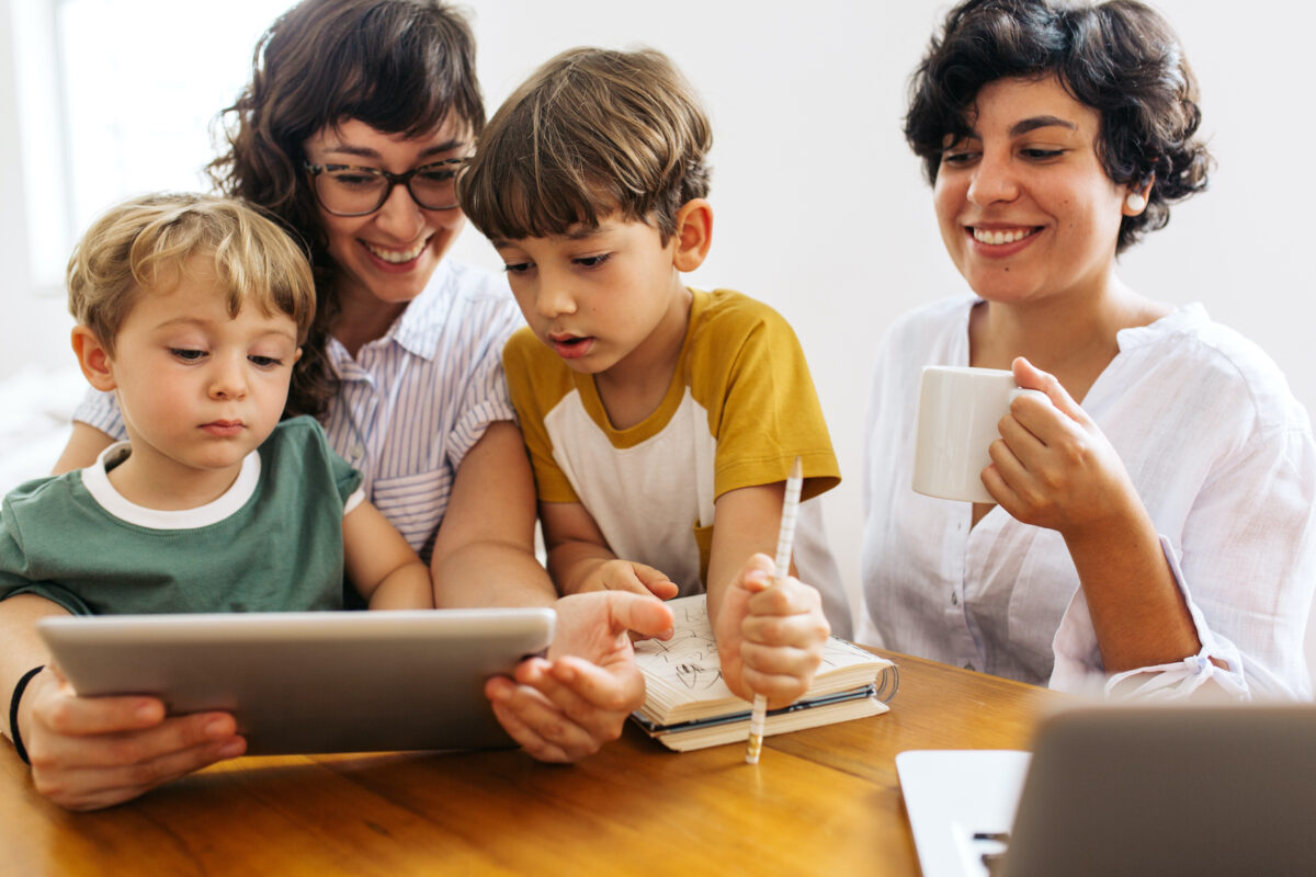 Family sitting together at table using digital tablet. Woman with kids looking at laptop with her partner drinking coffee and smiling.